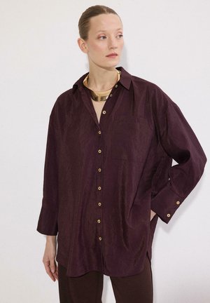 Žena so svetlou pokožkou a vlasmi stiahnutými dozadu má na sebe preveľkú tmavofialovú košeľu na gombíky a hrubý zlatý náhrdelník, pozerá sa bokom.