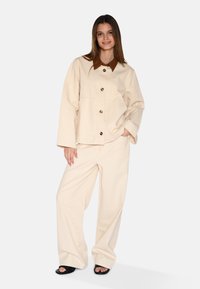 Beige oversized jas met bruin kraag, knoopsluiting en twee voorzakken, gecombineerd met bijpassende brede broek en zwarte sandalen.