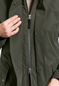 Groene waterdichte jas met een ritssluiting aan de voorkant, drukknoopsluitingen en een gladde textuur. Hand gepositioneerd nabij de rits voor detail.