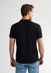 Černé polo tričko s texturovaným designem, krátkým rukávem a klasickým límcem, v kombinaci s světle modrými džíny. Pohled je zezadu.