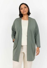 Cardigan lungo con apertura frontale in morbido tessuto verde oliva, maniche a tre quarti e tasche laterali, indossato sopra un top crema.