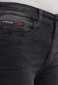 Svarta denimjeans har en liten bakficka med röda, vita och grå sömnadsdetaljer. Tyget har en slät textur och en lätt glans.