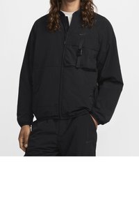 Černá bunda Nike s předním zipem, elastickými manžetami, kapsou na hrudi se stahovacím páskem a lehkou texturou.
