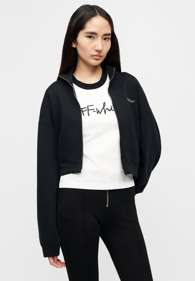 Μαύρο crop jacket με φερμουάρ, μείγμα βαμβακιού, με υπερμεγέθη μανίκια. Λευκό T-shirt από κάτω με την επιγραφή "OFF-white" σε μαύρο γράψιμο. Μαύρο στενό παντελόνι.