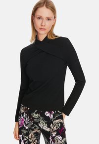 Schwarzes Langarmtop mit tailliertem Schnitt und gedrehtem Halsauschnitt. Kombiniert mit floral gemusterten Hosen, die den Kontrast und die Textur betonen.