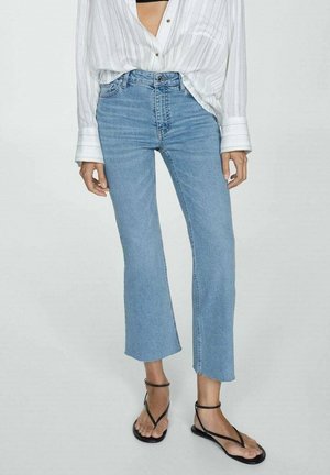 Vrouw draagt lichtblauwe cropped flare jeans, wit gestreept blouse met knopen en zwarte sandalen met bandjes, staand tegen een effen achtergrond.