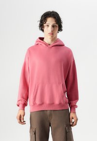 Hoodie en coton rose avec une poche avant, style kangourou, capuche avec cordon de serrage et poignets côtelés. Le modèle le porte avec un pantalon cargo marron.