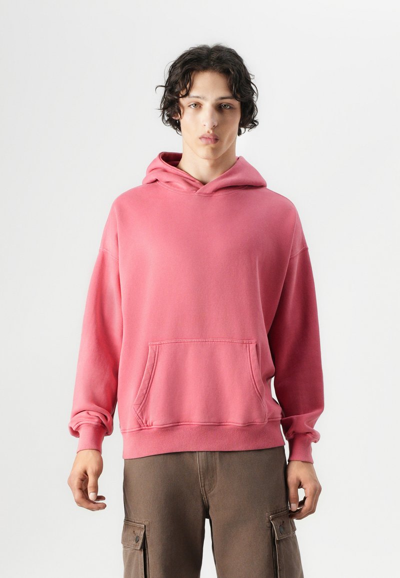 Hoodie en coton rose avec une poche avant, style kangourou, capuche avec cordon de serrage et poignets côtelés. Le modèle le porte avec un pantalon cargo marron.