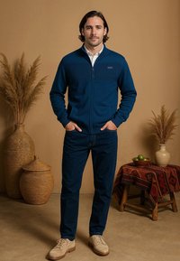 Marineblauwe ritsjack met horizontale geribbelde textuur, gecombineerd met donkerdenim broek en beige schoenen. Geplaatst tegen een neutrale achtergrond met decor.