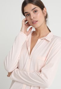 Camicia a maniche lunghe color rosa chiaro con colletto, chiusura a bottoni e una tasca sul petto. Il tessuto sembra morbido e liscio con una vestibilità comoda.