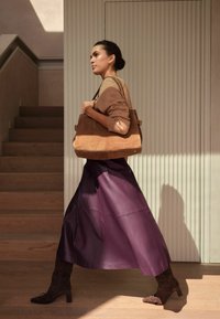 Bolso de ante marrón que sostiene una mujer con un suéter color beige y una falda de cuero púrpura, caminando junto a paneles de pared blancos texturizados y escaleras.