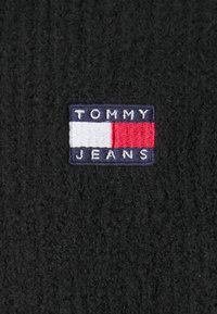 A fekete szövet szövetmintázattal rendelkezik, amelyen egy téglalap alakú hímzett folt található. A folton a "TOMMY JEANS" felirat és egy piros, fehér és tengerészkék színkombináció látható.