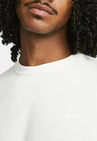 Vit bomullströja med rund halsringning, med en diskret, reflekterande Nike swoosh-logotyp på bröstet. Slät textur.