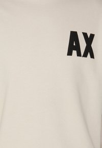T-shirt di colore crema realizzato in un tessuto morbido, con un logo ricamato nero "AX" in un carattere audace e moderno sul lato sinistro del petto.