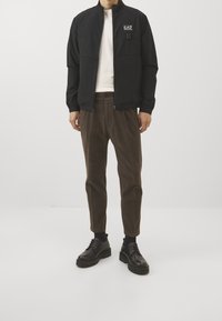 Svart zip-up jacka med logotyp, kombinerad med bruna spetspartade byxor i kordfläsk och svarta snörskor. Enkel design och figurnära snitt.