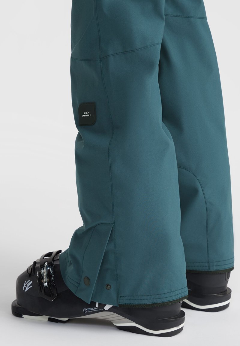 Teal Skihosen mit einer lockeren Passform, ausgestattet mit einem Logo-Patch, Knöpfen am Knöchel und wasserabweisendem Material, kombiniert mit schwarzen Skischuhen.