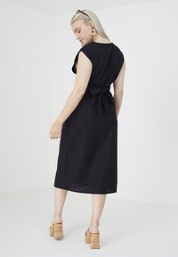 Abito midi nero con corpino aderente e dettaglio di legatura sul retro. La modella indossa scarpe con zeppa in sughero. Tessuto leggero, design minimal.