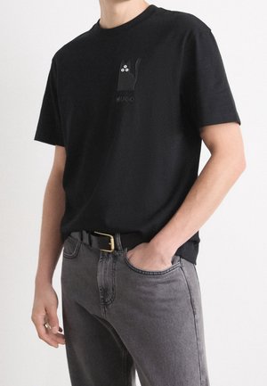 Homme portant un T-shirt noir avec un petit graphisme d'un chat noir et le mot « HUGO » sur la poitrine, associé à un jean gris et une ceinture noire.