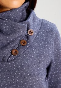 Modrý svetr s puntíky a rolákem, který má na límci tři hnědé knoflíky. Měkká tkanina s mírně texturovaným vzhledem.