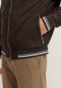 Braune Bomberjacke mit Reißverschluss vorne, Mesh-Textur, gerippte Bündchen mit weißen Streifen, getragen über beigen Hosen.