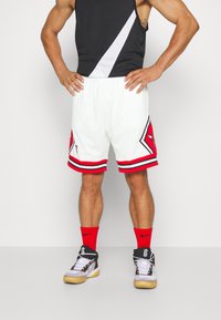 Pantalones cortos de baloncesto en malla blanca con detalles en rojo, con un logo en negro y rojo. Combinados con zapatillas en negro y blanco y calcetines rojos.