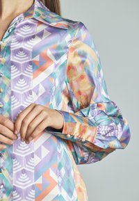 Camicia button-up colorata con motivi geometrici in viola, arancione e teal. Maniche lunghe con rifiniture plissettate e texture del tessuto liscia.