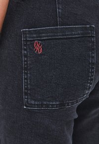 Tummansinisen farkut, joissa on takatasku punaisella brodeeratulla logolla. Kankaassa on sileä tekstuuri, jossa on hienovaraisia sävyvaihteluita.