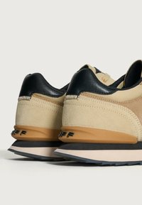 Sneaker che presentano un mix di suede beige e pelle nera, forma arrotondata, accenti arancioni prominenti e una suola testurizzata in marrone e nero.