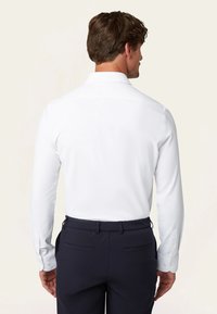 Weißes Langarmhemd mit ausgestelltem Kragen, ausgestattet mit Knopfmanschetten, getragen mit marineblauen Hosen. Glattes Material mit taillierter Passform.