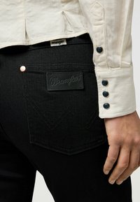 Jeans de denim negros con un parche de cuero de Wrangler en la parte trasera, que combinan botones de cobre y negros en una camisa de manga larga color crema.