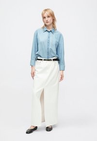 Camicia di jeans con colletto e dettagli sui bottoni blu, abbinata a una lunga gonna bianca con spacco laterale e cintura nera.