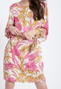 Blommig klänning med rosa och gula tropiska tryck på en krämfärgad bakgrund, med långa ärmar och en lös passform. Tyg med struktur och rosetter vid ärmsluten.