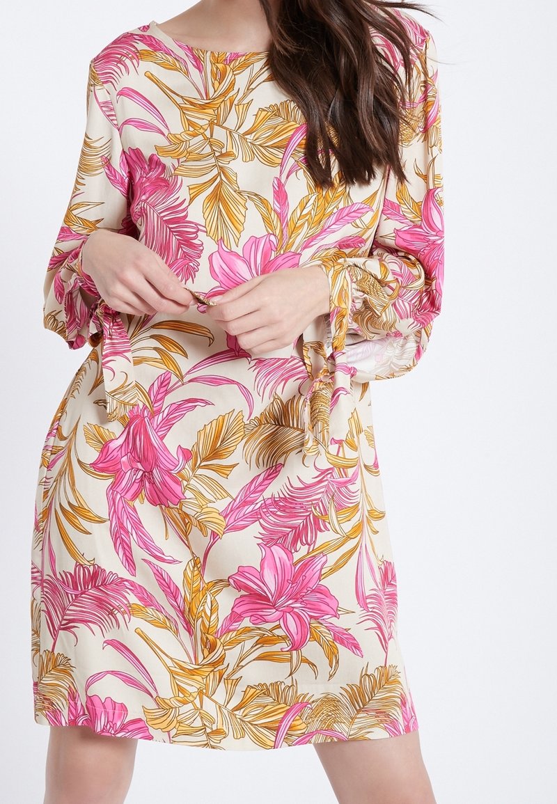 Blommig klänning med rosa och gula tropiska tryck på en krämfärgad bakgrund, med långa ärmar och en lös passform. Tyg med struktur och rosetter vid ärmsluten.