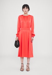 Langärmliges Satin-Kleid in lebhaftem Korallenrot, mit gerafftem Ausschnitt, elastischem Bund und einem schwingenden Midikleid. Schwarze Clutch und Riemchensandalen.