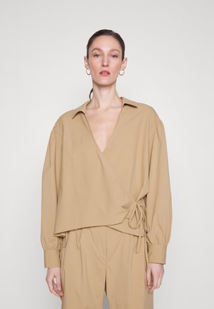 Blusa avvolgente color beige con scollatura a V profonda, maniche lunghe a palloncino, polsini elasticizzati e dettaglio annodato in vita; texture in cotone morbido.