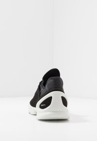 Musta mokkanahkainen ja nahkainen sneaker, jossa on valkoinen kumipohja, pyöristetty muoto ja kontrastiset tekstuurikoristeet kantapäässä.