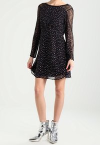 Schwarzes Blumenkleid mit langen Spitzenärmeln, tailliertem Bund und fließendem Rock. Kombiniert mit glänzenden silbernen Stiefeletten.