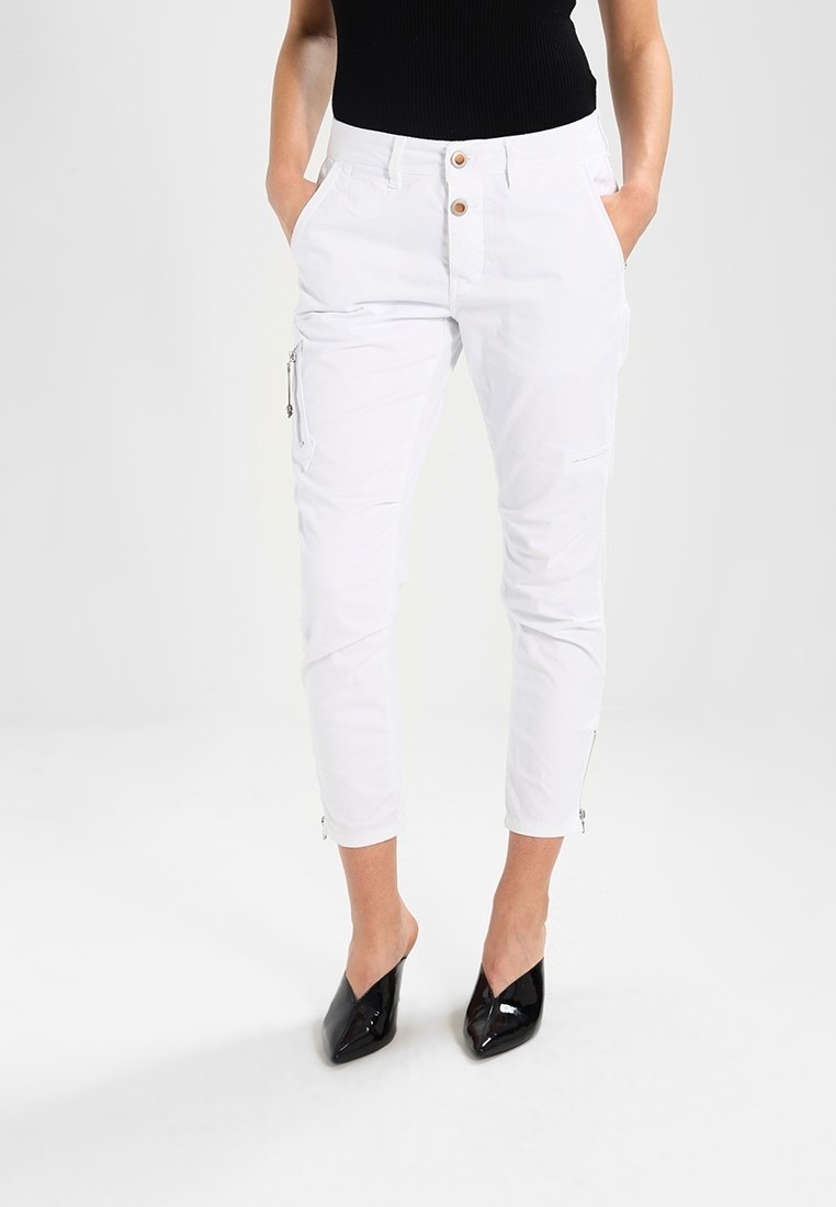 Femme portant un pantalon blanc court avec poche zippée latérale et talons noirs pointus, mains dans les poches, sur fond uni.