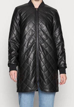 Musta tikattu pitkä takki, jossa edessä vetoketju ja resorireunaiset hihansuut, pukeutuneena henkilö, jolla on vaaleansiniset farkut ja yksivärinen tausta.