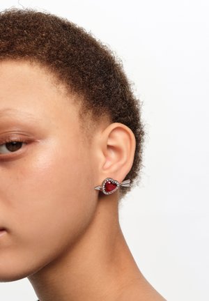 Primer plano de una oreja con un pendiente de plata en forma de corazón rojo atravesado por una flecha, con pequeñas piedras transparentes alrededor del corazón.