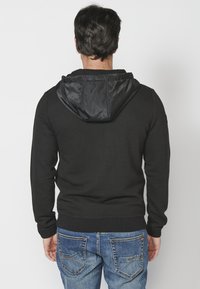 Hoodie nero in tessuto strutturato con cappuccio liscio a contrasto, polsini e orlo a costine; abbinato a jeans blu con tasche posteriori distintive.