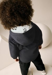 Veste marine avec capuche, dotée d'une doublure bleu clair affichant des visages souriants. Un pantalon noir et des baskets blanches complètent l'ensemble.