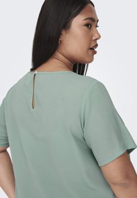 Top a maniche corte, verde chiaro, realizzato in tessuto morbido. Presenta uno scollo a chiave sul retro con chiusura a bottone e un collo rotondo.