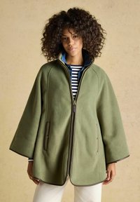 Frau mit lockigem Haar, die einen olivgrünen Cape-Mantel mit Reißverschluss über einem gestreiften Hemd und weißen Hosen trägt, stehend vor einem beigefarbenen Hintergrund.