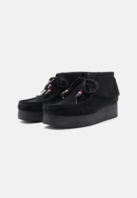 Botas de tornozelo em camurça preta com um design com franjas, laços decorativos com contas e uma sola grossa e texturizada para maior altura e aderência.