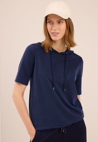 Žena v námořnickém krátkém rukávu s kapucí a béžovou čepicí, pózující s jednou rukou v kapse na světlém pozadí.
