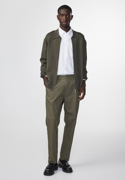 Veste zippée vert olive, chemise blanche à boutons, pantalon olive et chaussures en cuir noires. Textures lisses et design décontracté.