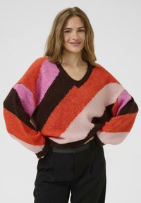 Pull en V avec des rayures diagonales audacieuses en orange, rose, marron et rose clair. Fabriqué en tissu texturé avec des manches surdimensionnées.
