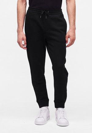 Mann trägt schwarze Jogginghose mit Kordelzug und weiße Sneakers, steht vor einem schlichten weißen Hintergrund.