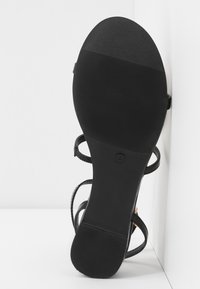 Sandale noire avec une semelle texturée, dotée de fines lanières et d'une boucle sécurisée. La semelle est plate avec une légère courbure pour le confort.